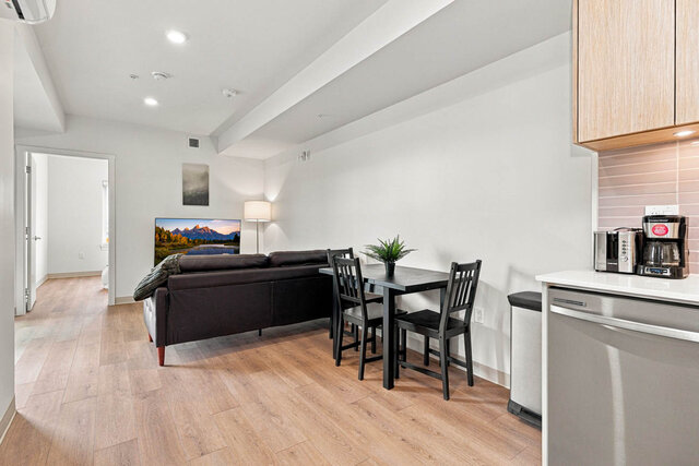 Dining table with four chairs beside the kitchen, next to the living area with a leather sofa, TV and floor lamp.