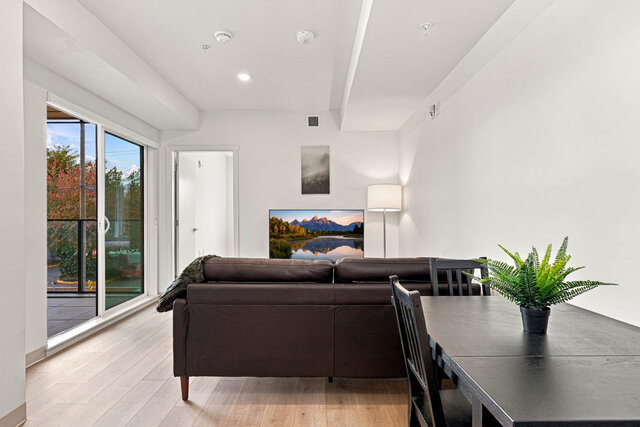 Living room with a leather sofa facing the TV, a dining table and a sliding glass door to the balcony. - 213-1529 Oxford Street, North Vancouver