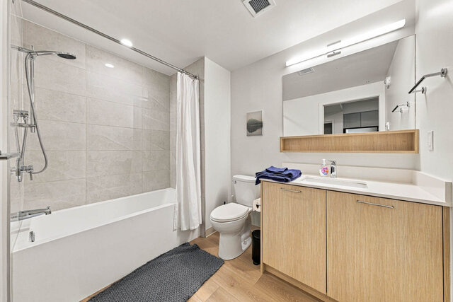 Second full bathroom with a tub and shower, a tile surround and a light wood vanity under a wide mirror and open shelf.
