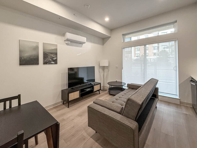 Living area with a grey tufted sofa facing a TV on a media console, beside a sliding door with a transom window above.