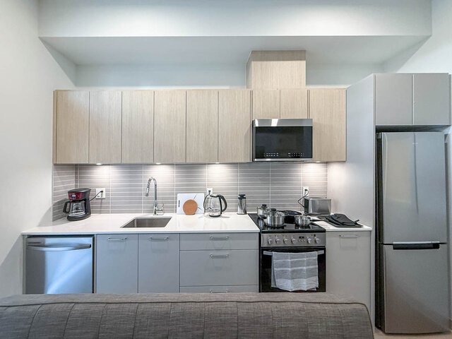 Kitchen along one wall with light oak upper cabinets, under-cabinet lights, quartz counters, a tile backsplash and stainless 