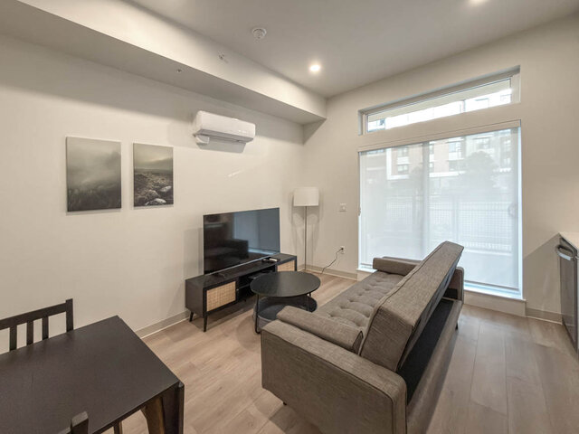 Living area with a grey tufted sofa, TV and coffee table, and a tall sliding door with a transom window. - 101-1529 Oxford Street, North Vancouver