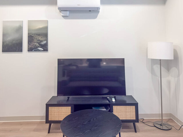 TV on a media console with a round coffee table, floor lamp and art under the air conditioner. - 101-1529 Oxford Street, North Vancouver