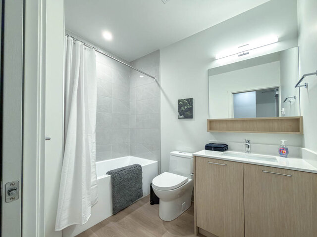 Full bathroom with a tub and shower, tile surround, a wood vanity with a quartz counter and a lit mirror with an open shelf.