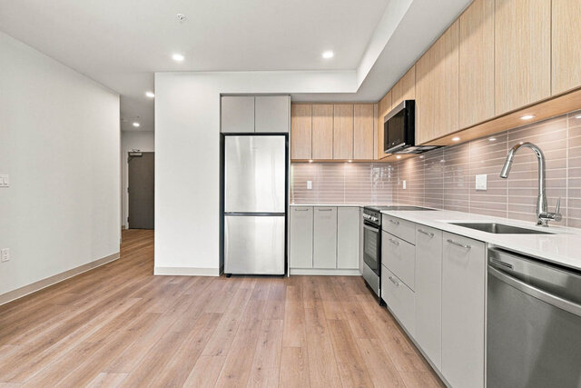 Kitchen corner with light oak upper cabinets, a tile backsplash and stainless appliances, with the hall to the front door beh