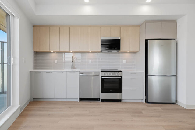 Straight-on view of the kitchen with light oak upper cabinets, quartz counters, a tile backsplash and stainless fridge, range