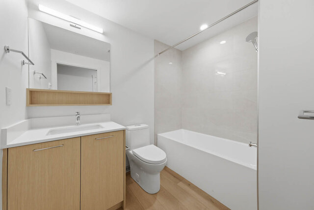 Second full bathroom with a tub and shower, tile surround, a wood vanity with a quartz counter and a lit mirror.