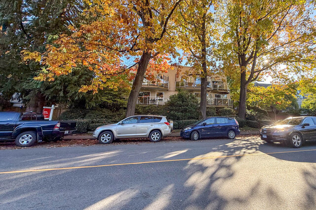 Low-rise building with glass-railed balconies behind tall autumn trees and hedges, seen from across the street.