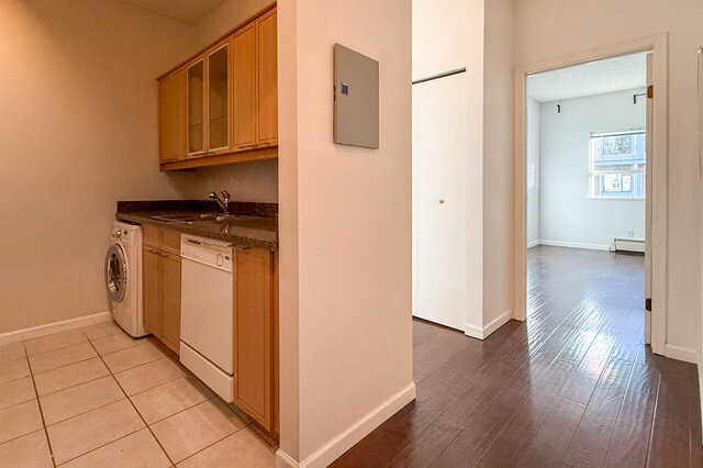 Hallway with the kitchen counter, washer-dryer and dishwasher on one side and the open bedroom door ahead, hardwood floors.