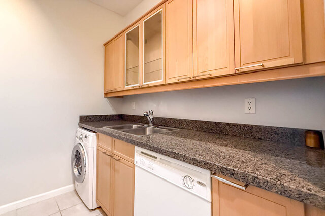 Kitchen counter with granite countertops, a double sink, glass-front wood cabinets, a dishwasher and a washer-dryer.