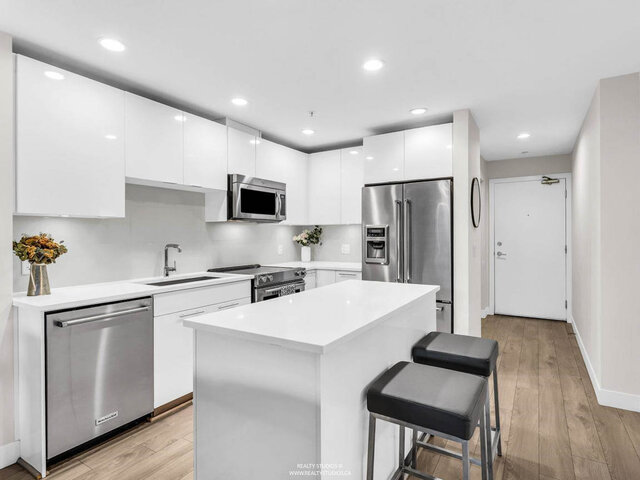 Kitchen with white gloss cabinets, white counters, stainless steel appliances and an island with two stools; the front door a