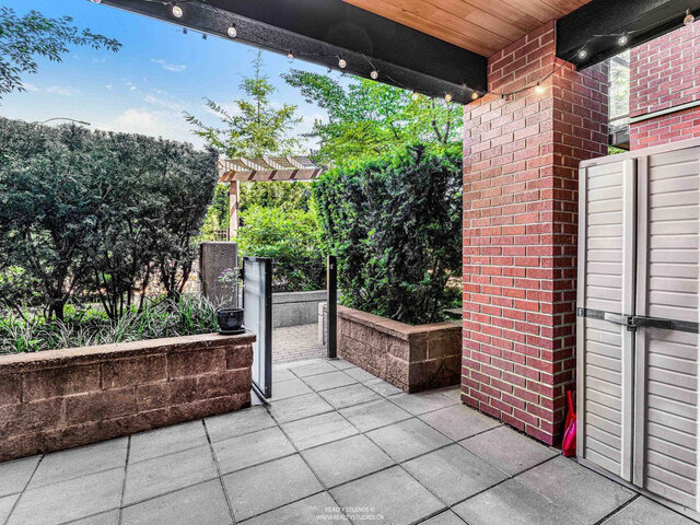 Private covered patio with stone pavers, string lights, a storage cabinet and a gate opening to the garden path, edged by hed