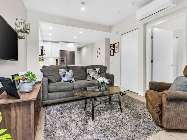 Living room with a grey sofa and glass coffee table, the kitchen behind it and a closet and air conditioning unit to the righ