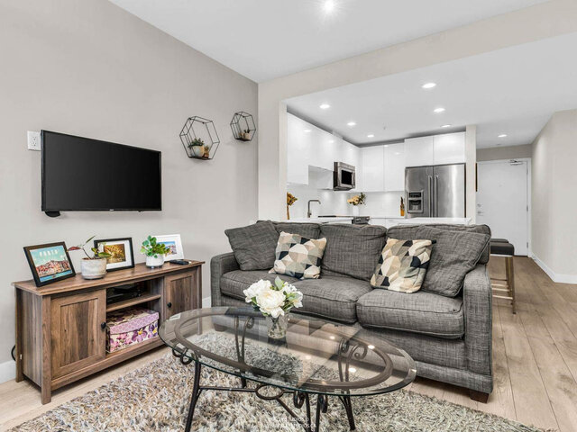 Living room with a wall-mounted TV and sofa, opening to the white kitchen and the front door. - 117-723 3rd street West, North Vancouver