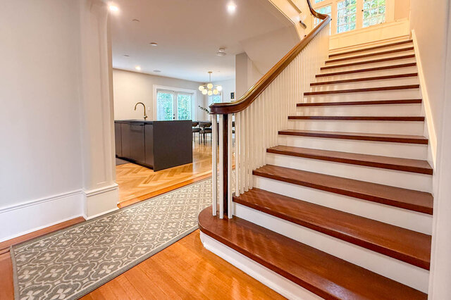 Grand staircase with a wood handrail and white spindles, beside the kitchen with herringbone floors. - North Vancouver