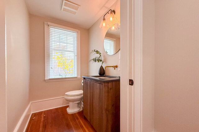 Main-floor powder room with a wood vanity, a wall-mounted brass faucet and a round mirror. - North Vancouver