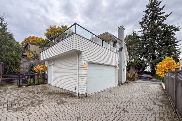 Garage with the glass-railed deck above, a paver driveway and the gated yard beside it. - North Vancouver